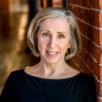 A smiling woman with chin-length hair and blue eyes smiles, standing next to a brick wall.