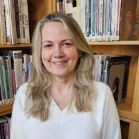 A smiling woman with shoulder length blonde hair stands in front of a bookshelf.