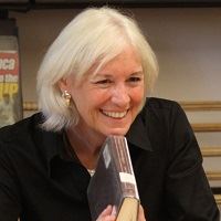 A smiling woman with chin-length white hair and gold earrings, holding a book.